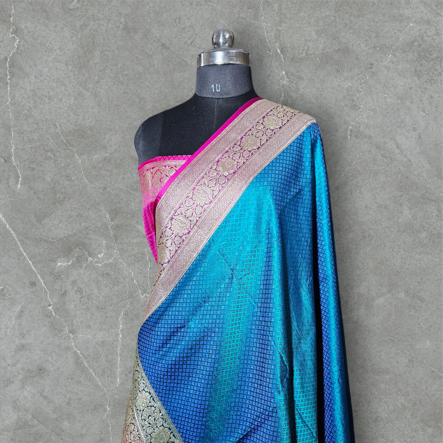 A luxurious Banarasi silk saree displayed on a mannequin, featuring a rich combination of pink, blue, and gold tones. The saree is adorned with intricate gold zari weaving in traditional floral and geometric patterns, with a broad golden border adding elegance. The vibrant color gradient enhances its beauty, making it perfect for festive and wedding occasions.