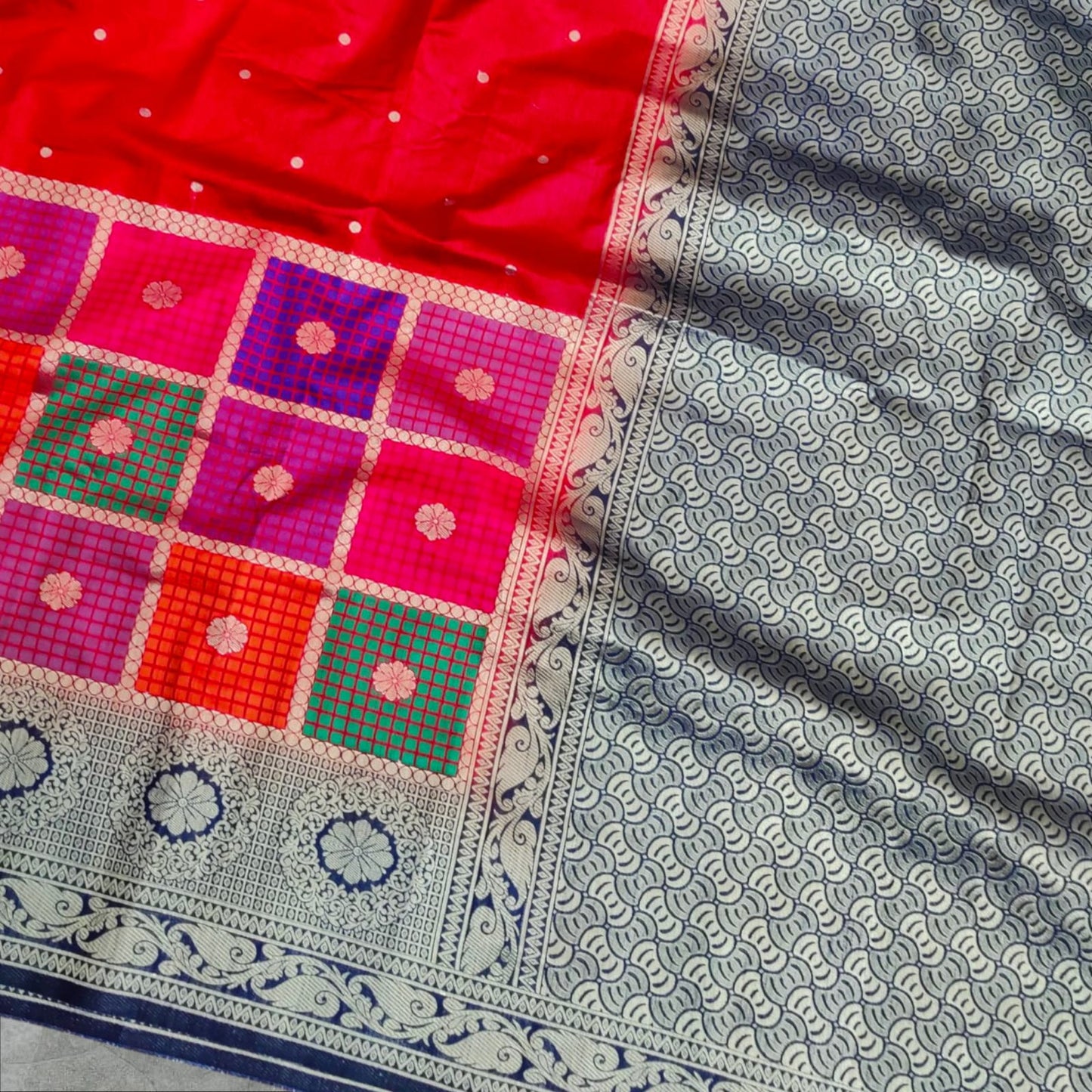 A vibrant saree with a striking combination of colors and intricate patterns. The upper part features a rich red base with small white dots, followed by a checkered design in shades of pink, purple, orange, and green, each square adorned with delicate floral motifs. The lower part and pallu contrast beautifully with a gray and navy blue intricate wave-like pattern, bordered with elegant floral and vine motifs. The saree blends traditional and contemporary aesthetics,