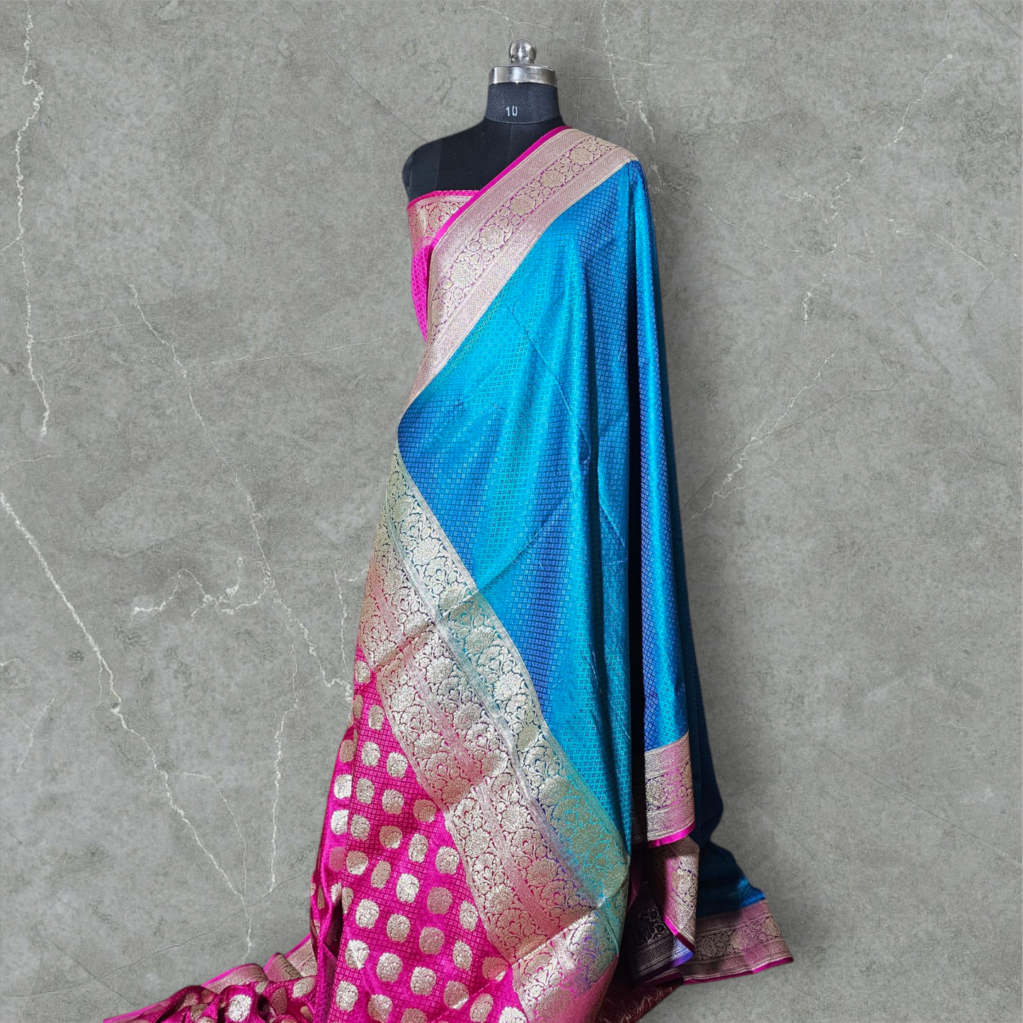A luxurious Banarasi silk saree displayed on a mannequin, featuring a rich combination of pink, blue, and gold tones. The saree is adorned with intricate gold zari weaving in traditional floral and geometric patterns, with a broad golden border adding elegance. The vibrant color gradient enhances its beauty, making it perfect for festive and wedding occasions.