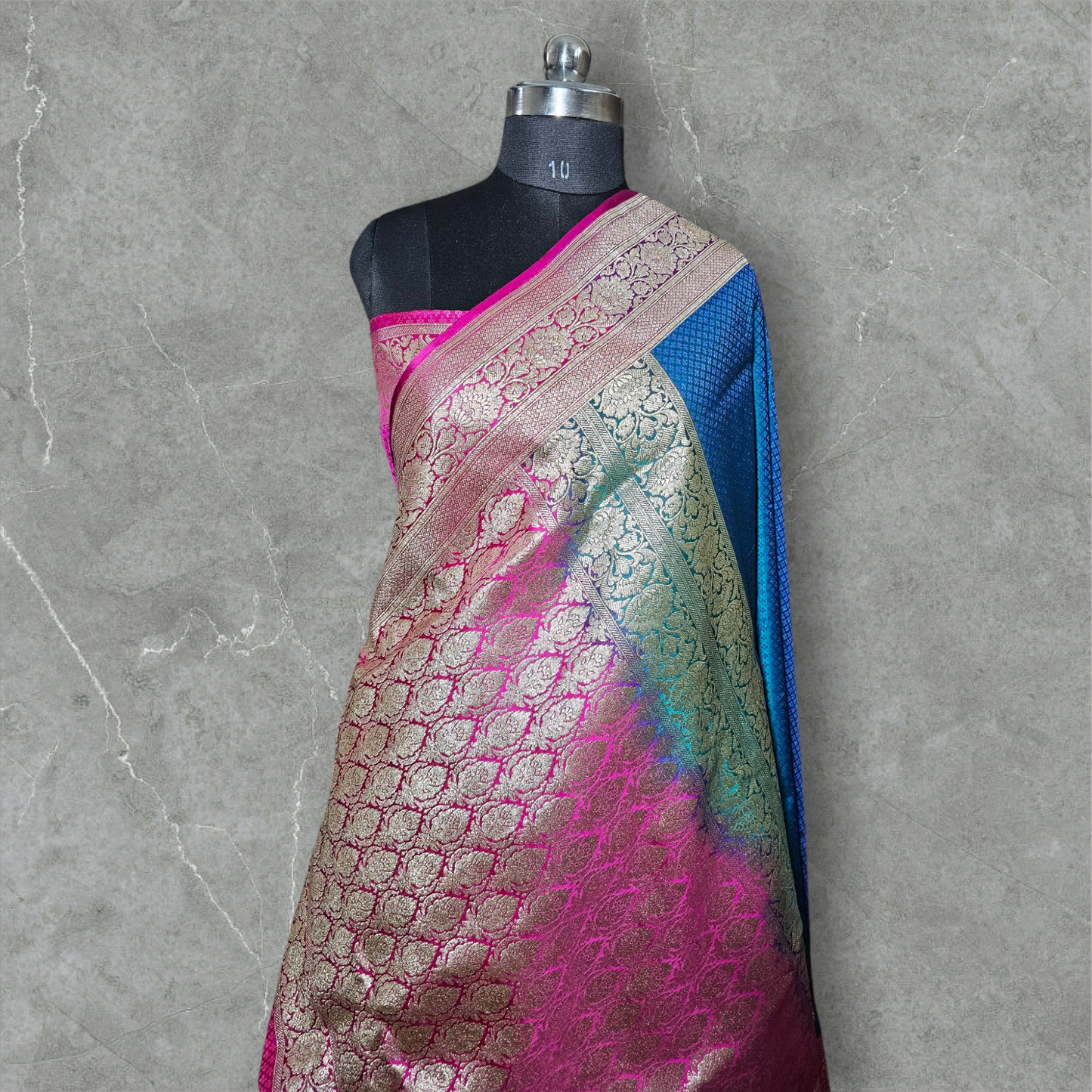 A luxurious Banarasi silk saree displayed on a mannequin, featuring a rich combination of pink, blue, and gold tones. The saree is adorned with intricate gold zari weaving in traditional floral and geometric patterns, with a broad golden border adding elegance. The vibrant color gradient enhances its beauty, making it perfect for festive and wedding occasions.