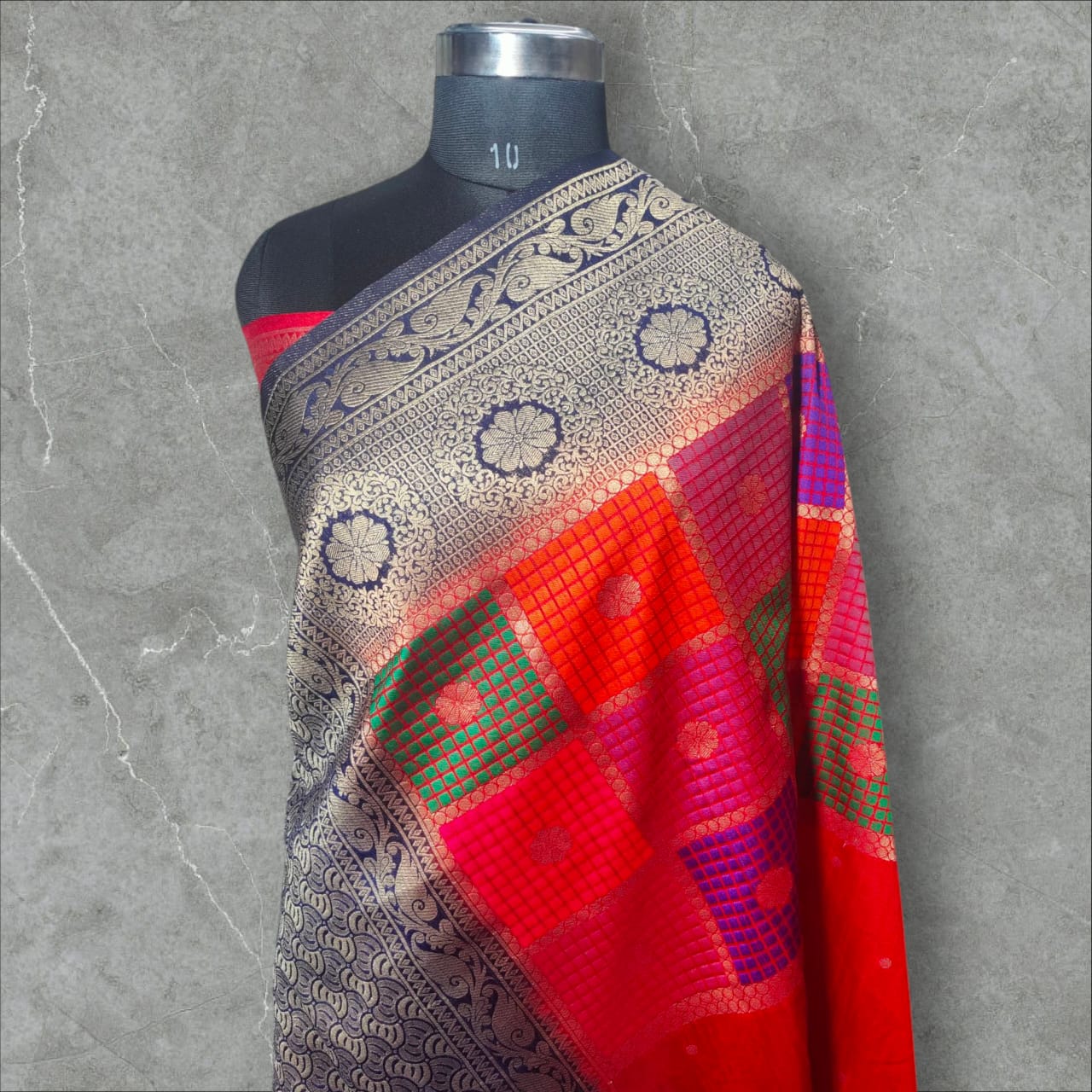 A vibrant saree with a striking combination of colors and intricate patterns. The upper part features a rich red base with small white dots, followed by a checkered design in shades of pink, purple, orange, and green, each square adorned with delicate floral motifs. The lower part and pallu contrast beautifully with a gray and navy blue intricate wave-like pattern, bordered with elegant floral and vine motifs. The saree blends traditional and contemporary aesthetics,