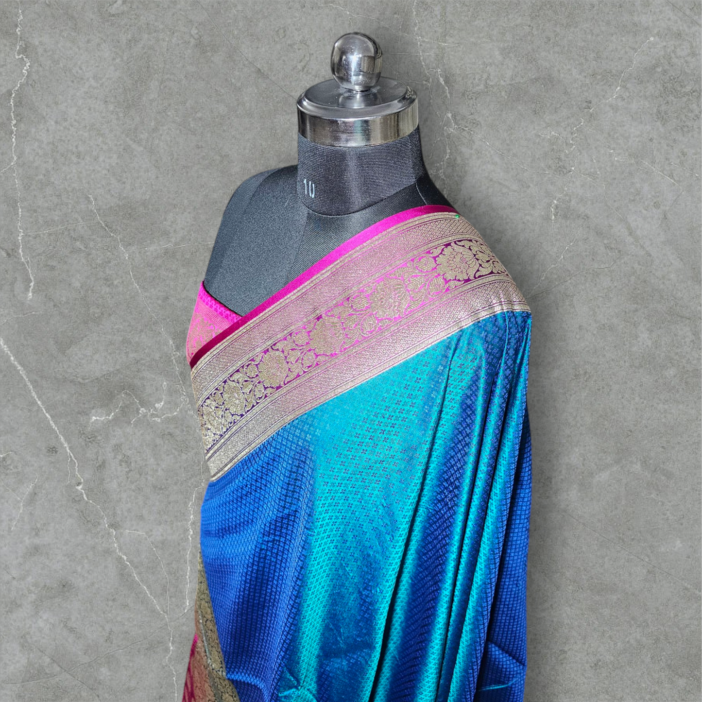 A luxurious Banarasi silk saree displayed on a mannequin, featuring a rich combination of pink, blue, and gold tones. The saree is adorned with intricate gold zari weaving in traditional floral and geometric patterns, with a broad golden border adding elegance. The vibrant color gradient enhances its beauty, making it perfect for festive and wedding occasions.
