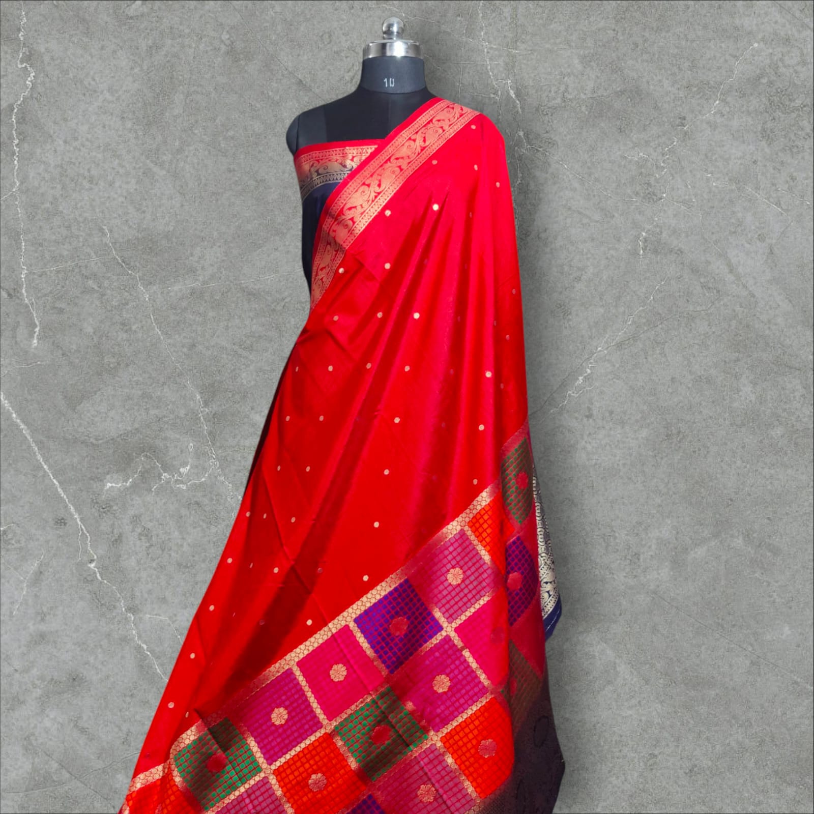 A vibrant saree with a striking combination of colors and intricate patterns. The upper part features a rich red base with small white dots, followed by a checkered design in shades of pink, purple, orange, and green, each square adorned with delicate floral motifs. The lower part and pallu contrast beautifully with a gray and navy blue intricate wave-like pattern, bordered with elegant floral and vine motifs. The saree blends traditional and contemporary aesthetics, making it perfect for festive 