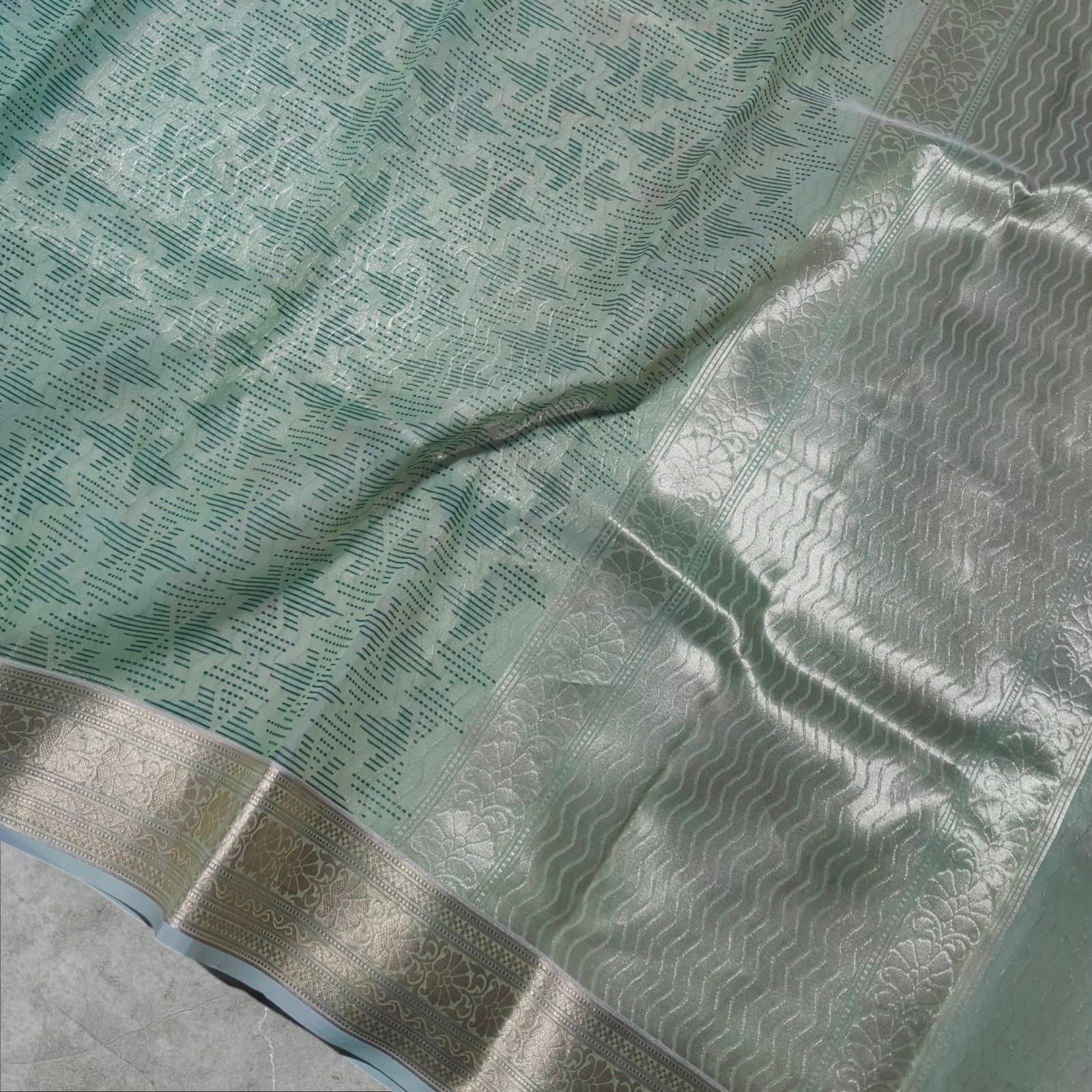 A graceful pastel green silk saree featuring intricate golden zari work on the border with floral and geometric patterns. The saree’s body has a delicate dotted and lined pattern, adding depth and elegance. Its soft sheen and rich golden accents make it a perfect choice for festive occasions, weddings, and cultural celebrations.