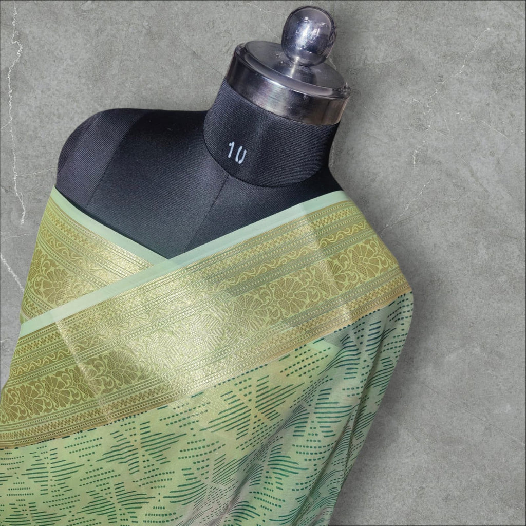 A graceful pastel green silk saree featuring intricate golden zari work on the border with floral and geometric patterns. The saree’s body has a delicate dotted and lined pattern, adding depth and elegance. Its soft sheen and rich golden accents make it a perfect choice for festive occasions, weddings, and cultural celebrations.