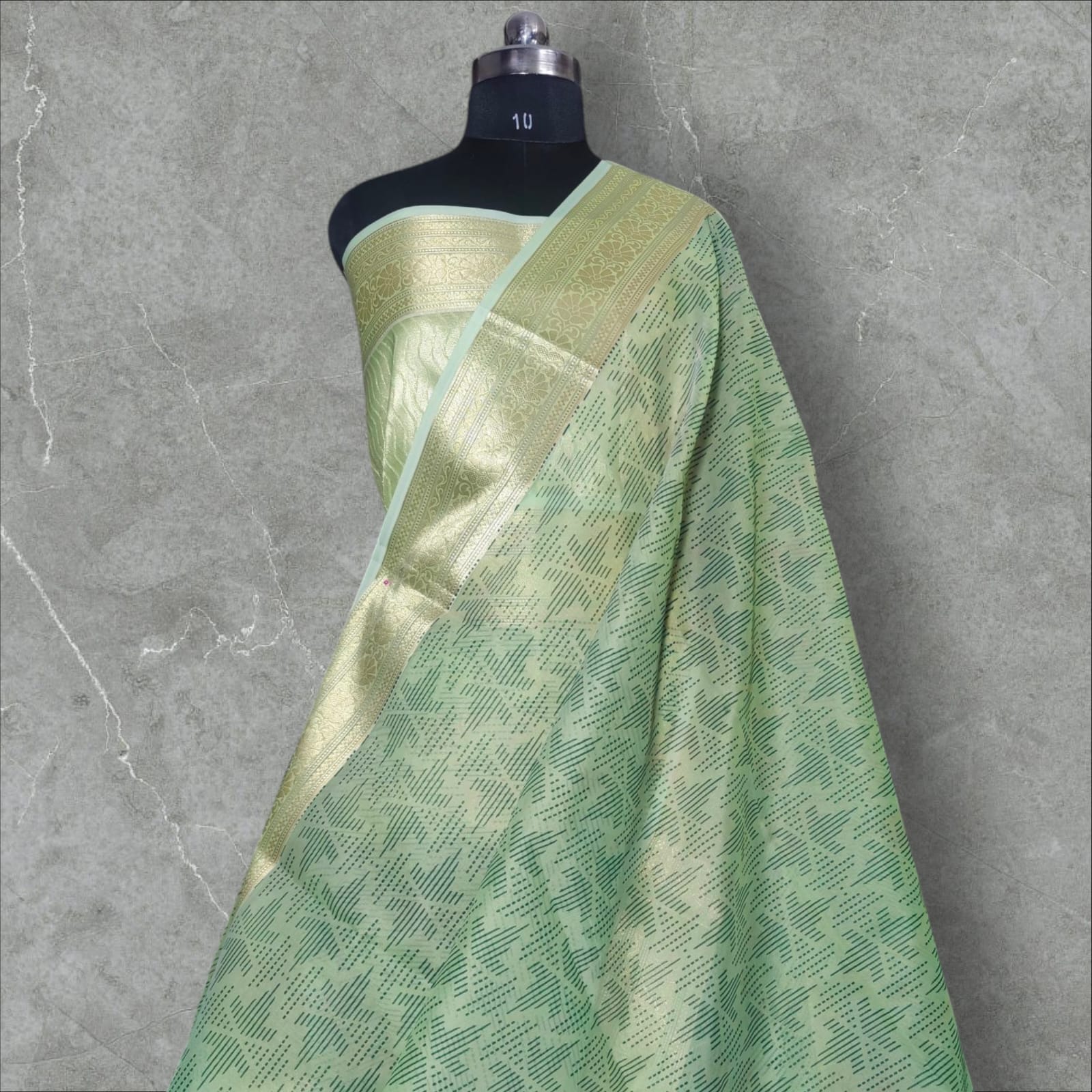 A graceful pastel green silk saree featuring intricate golden zari work on the border with floral and geometric patterns. The saree’s body has a delicate dotted and lined pattern, adding depth and elegance. Its soft sheen and rich golden accents make it a perfect choice for festive occasions, weddings, and cultural celebrations.