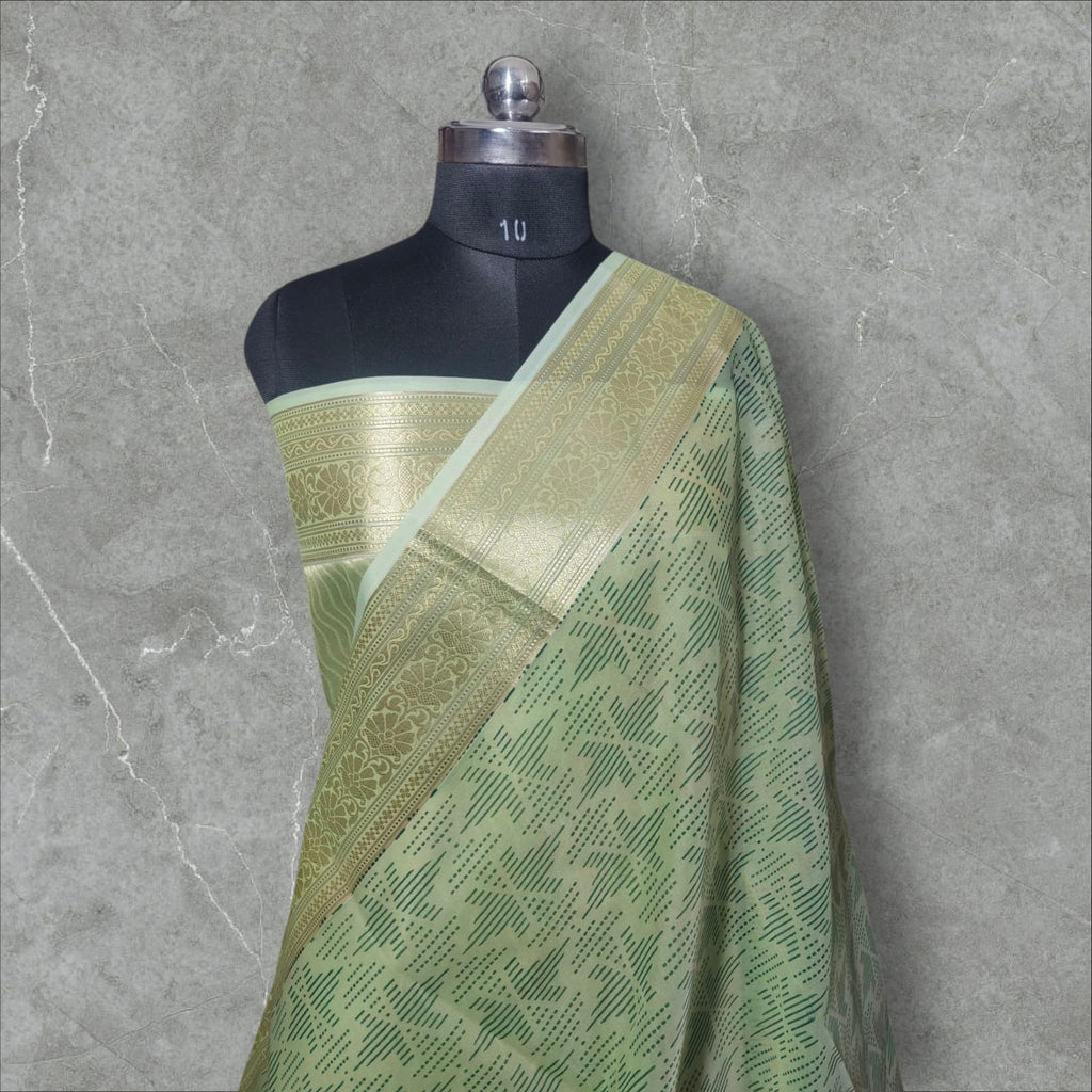 A graceful pastel green silk saree featuring intricate golden zari work on the border with floral and geometric patterns. The saree’s body has a delicate dotted and lined pattern, adding depth and elegance. Its soft sheen and rich golden accents make it a perfect choice for festive occasions, weddings, and cultural celebrations.